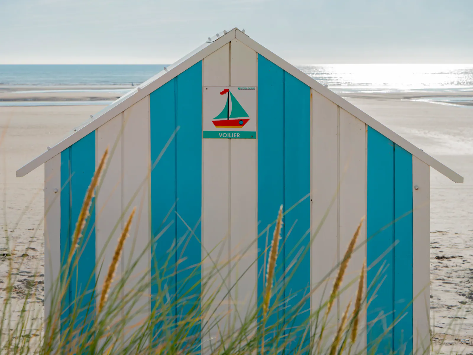 Cabine de plage traditionnelle bleue et blanche avec voilier peint, plage de sable fin en arrière-plan