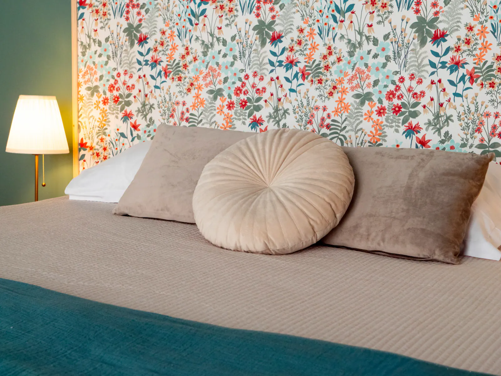 Chambre avec lit beige, coussins taupe, papier peint floral multicolore et lampadaire doré
