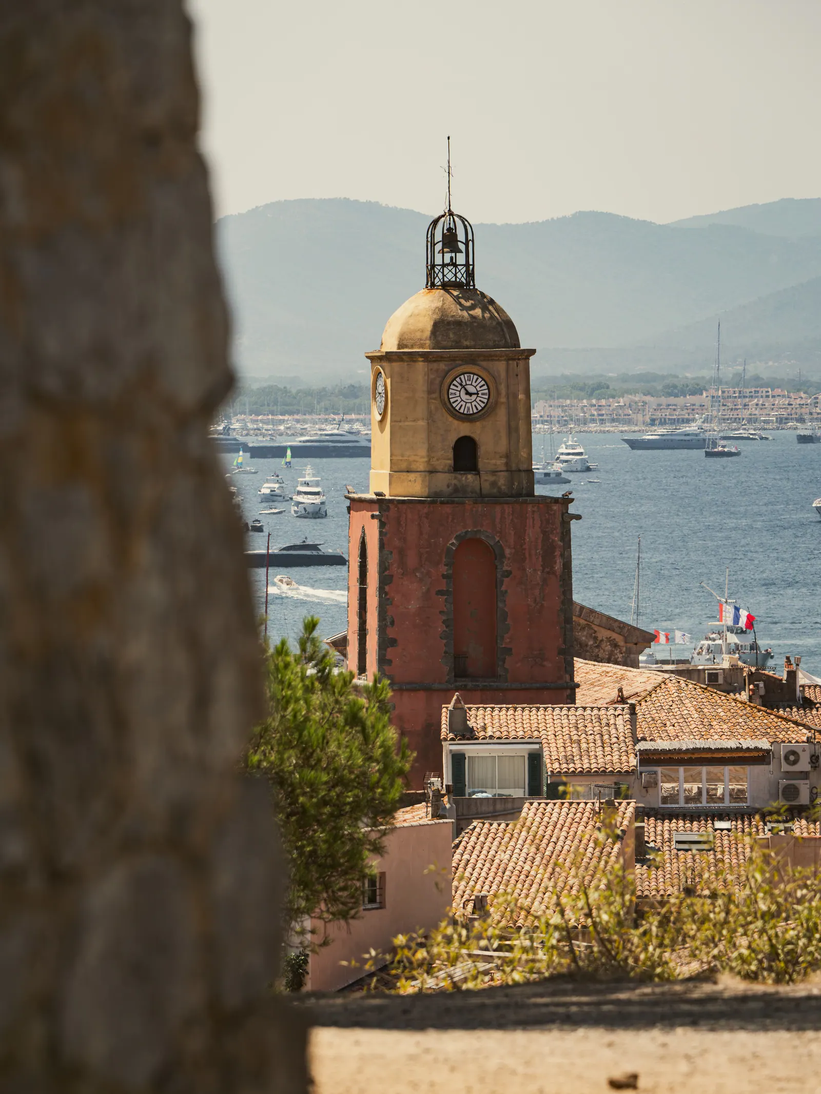 Clocher en pierre dominant le port de St-Tropez avec bateaux en arrière-plan et baies vitrées de la côte