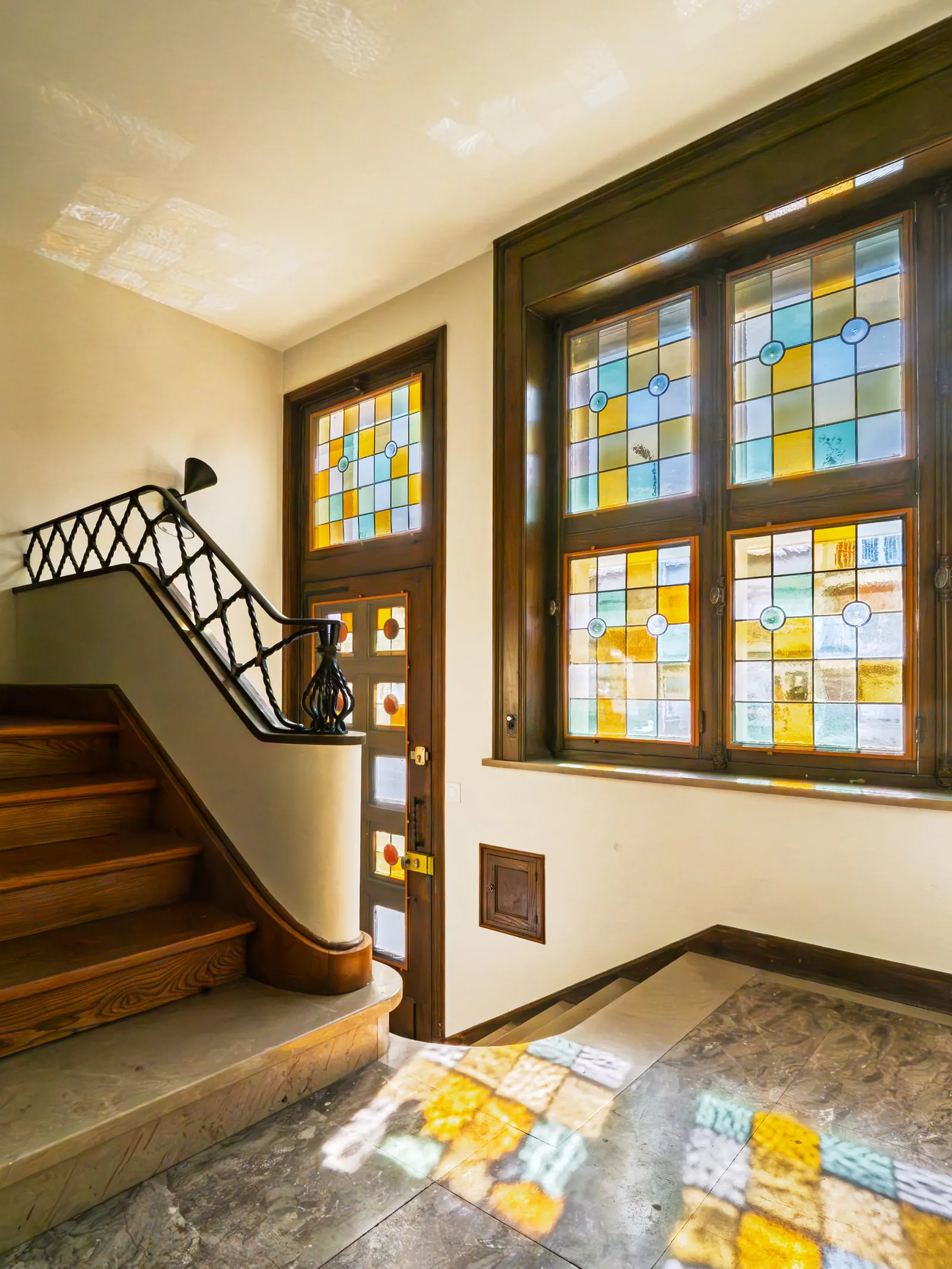 Escalier en bois avec rampe noire style art déco et vitraux jaunes bleus à motifs géométriques dans un hall d'entrée lumineux