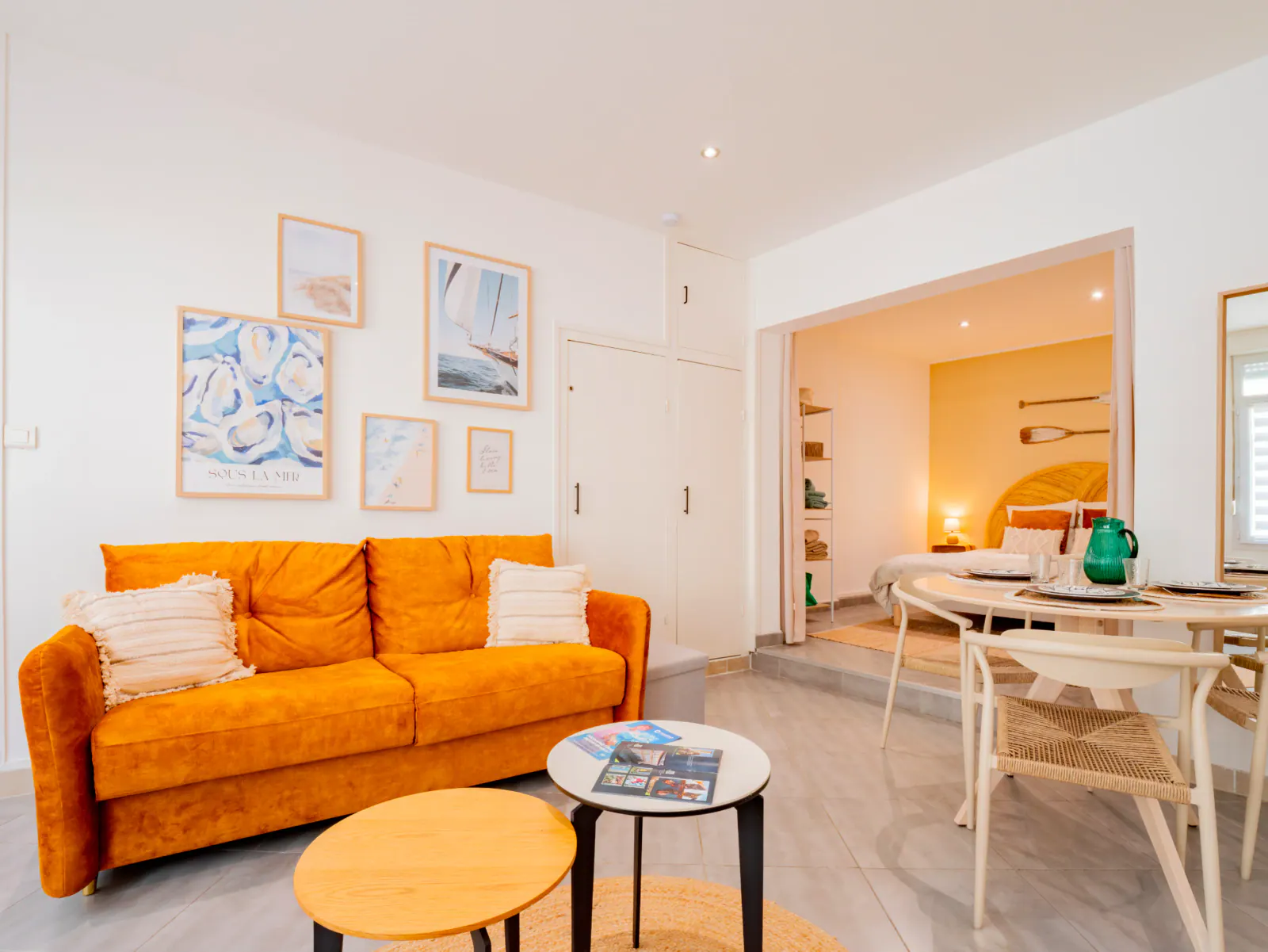 Salon lumineux d'un gîte côtier avec canapé ocre, tableaux marins et vue sur chambre