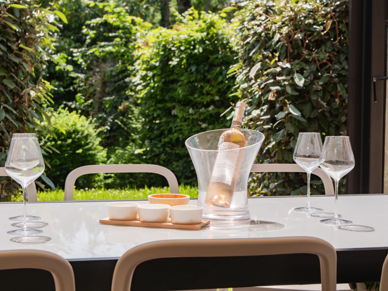 Table dressée en terrasse avec verres, bouteille de vin rosé et vue sur jardin luxuriant en arrière-plan